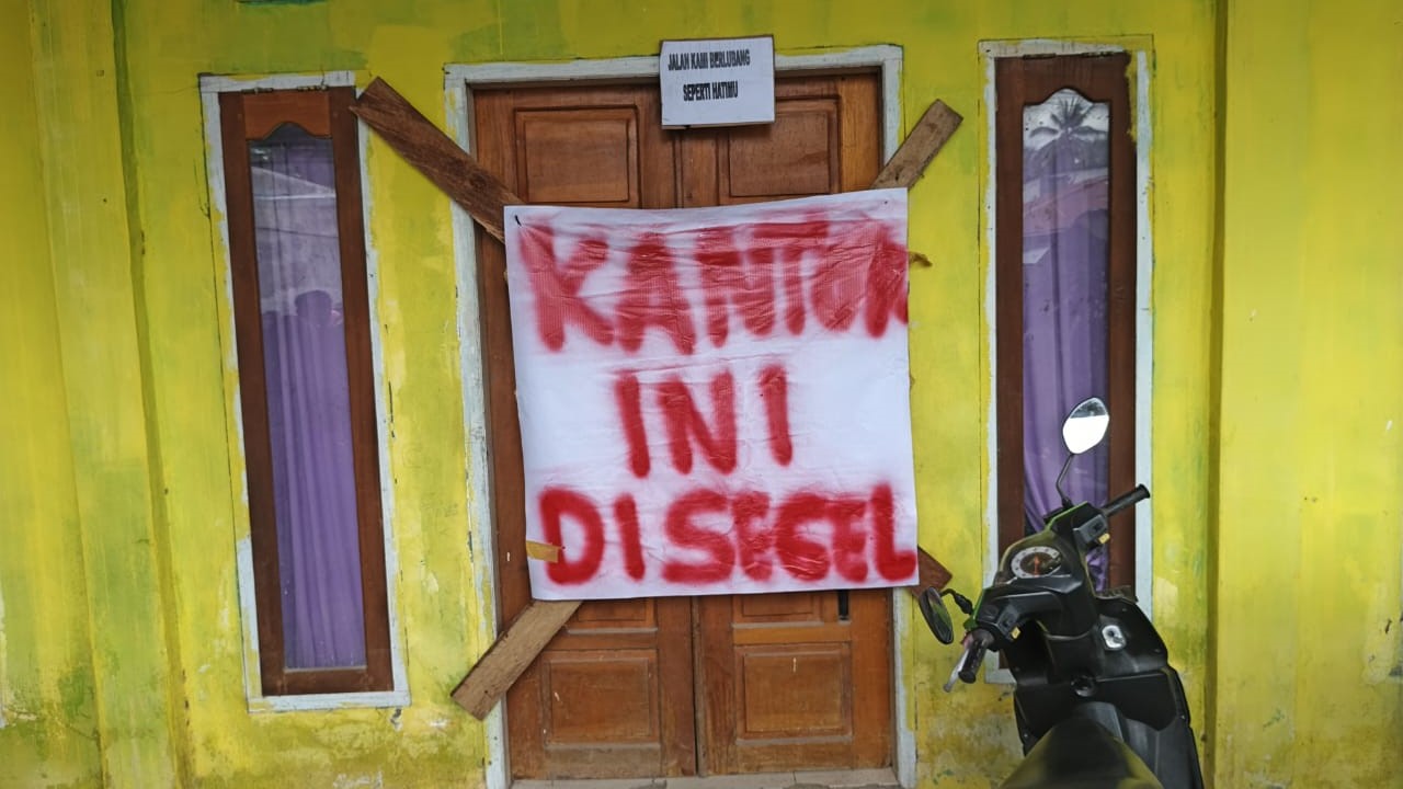 Pintu kantor Desa Torue dipalang dengan kayu dan dipasangi spanduk putih bertuliskan “KANTOR INI DISEGEL” dengan cat semprot merah.